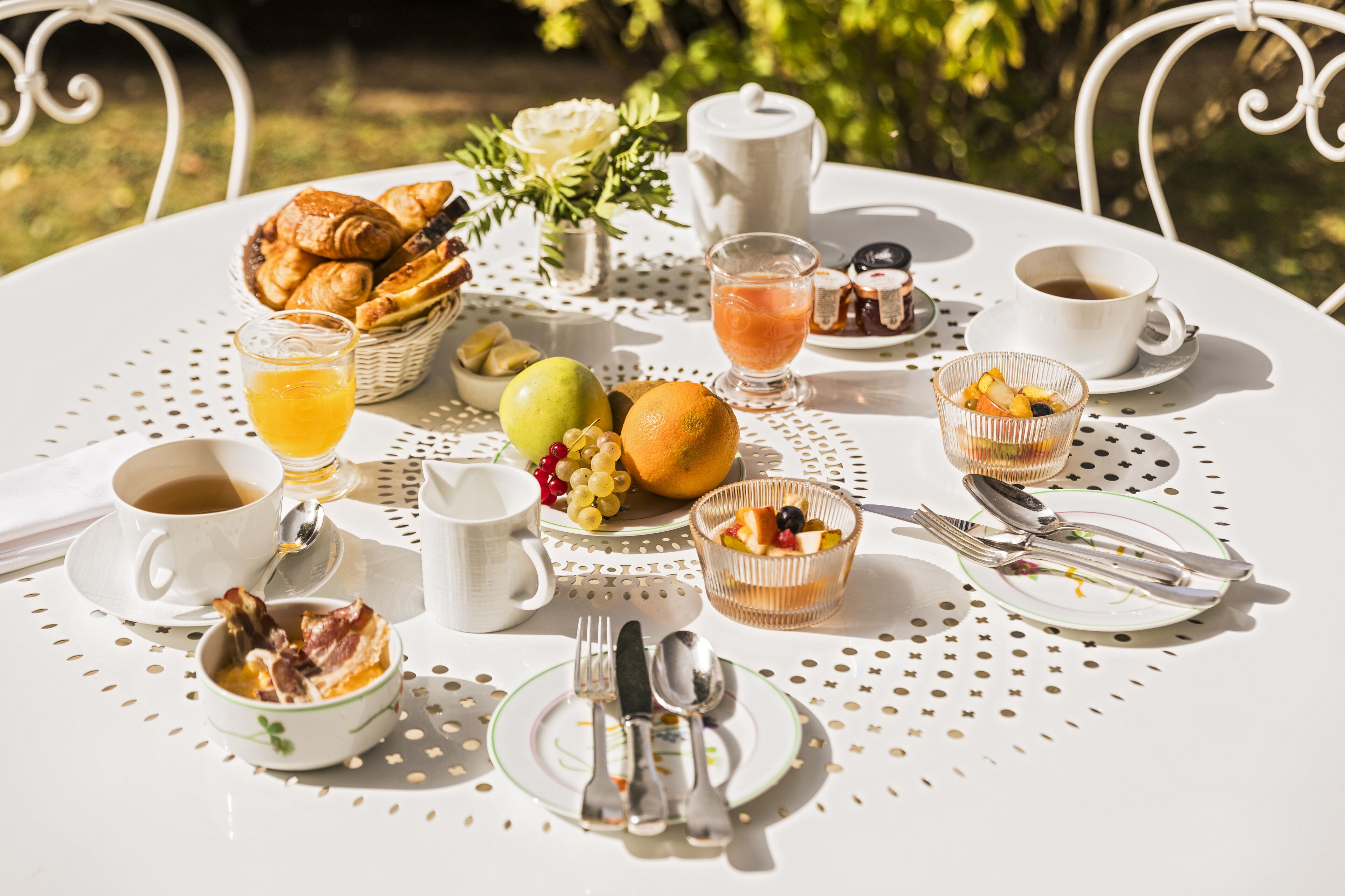 Mesa de pequeno-almoço posta com croissants, frutas, um bule, chávenas e utensílios.