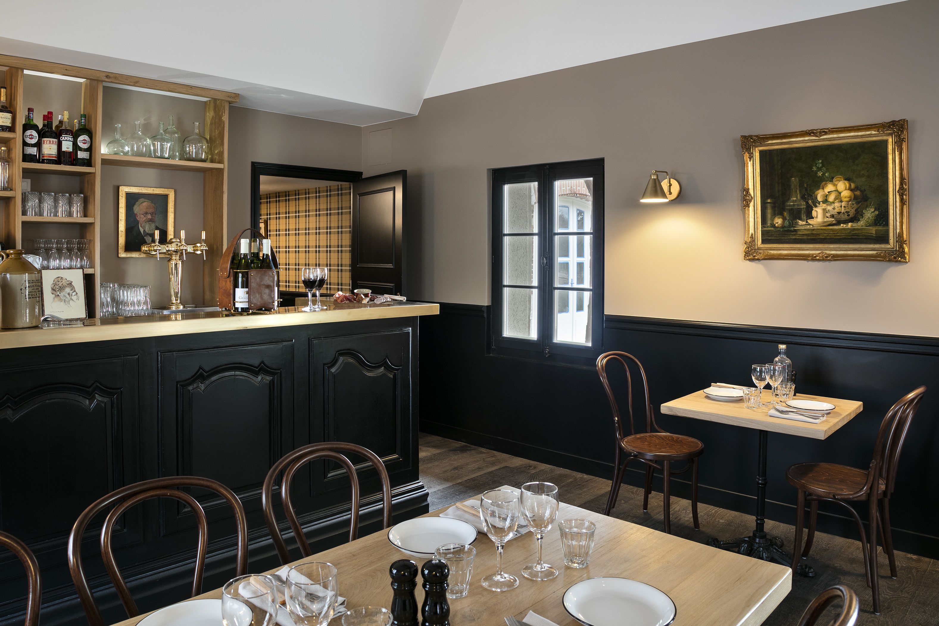 A dining area with a bar, wooden chairs, a table set for two, and a painting on the wall.