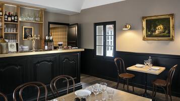 A dining area with a bar, wooden chairs, a table set for two, and a painting on the wall.