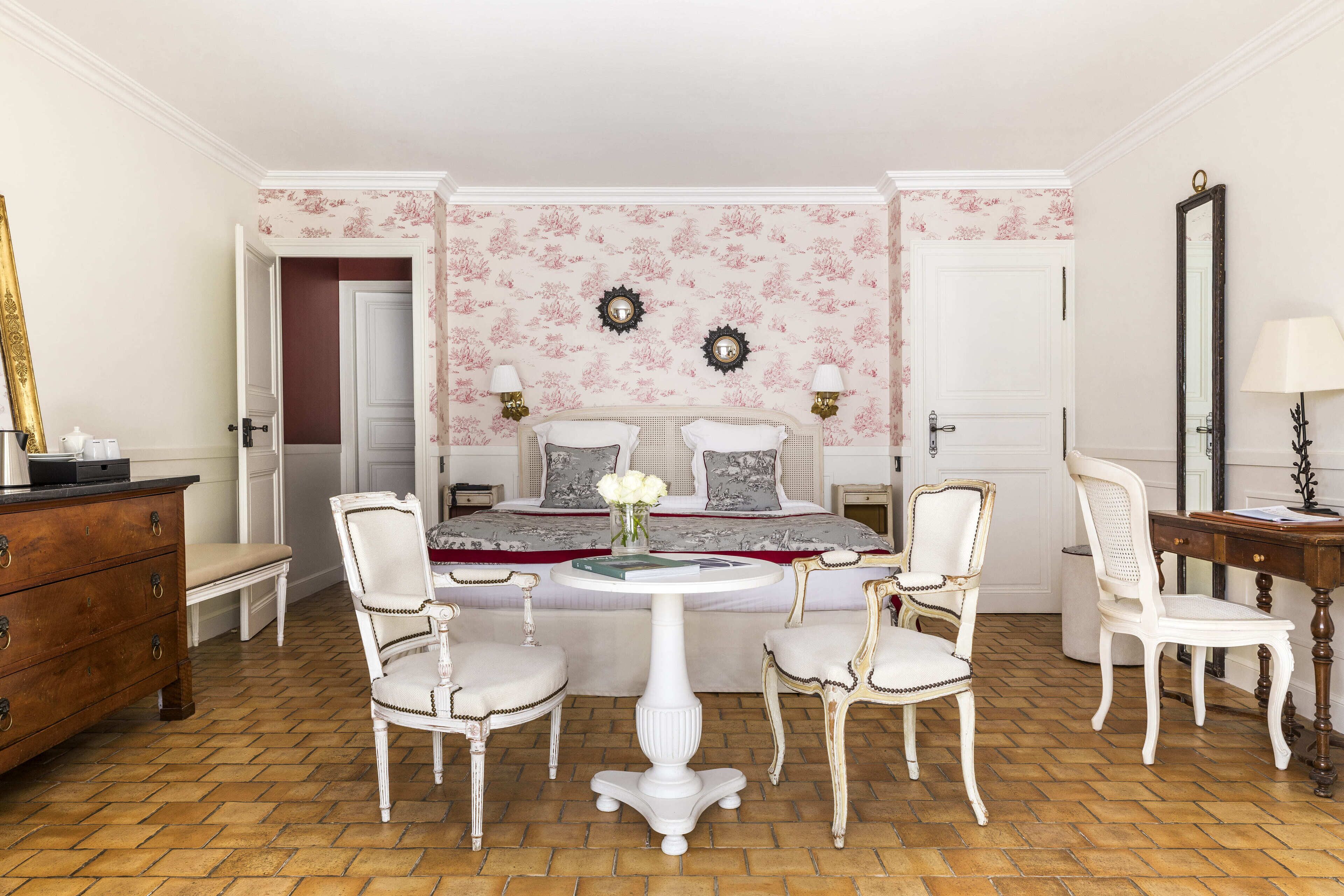A bedroom with a bed, two chairs, a dresser, a table, and a mirror.