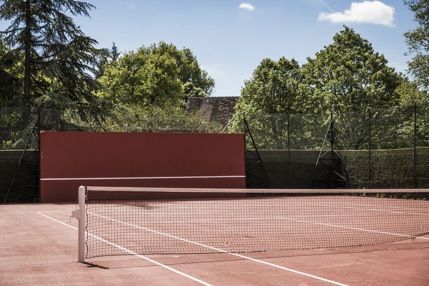 A tennis court with a net, surrounded by trees and a red wall.