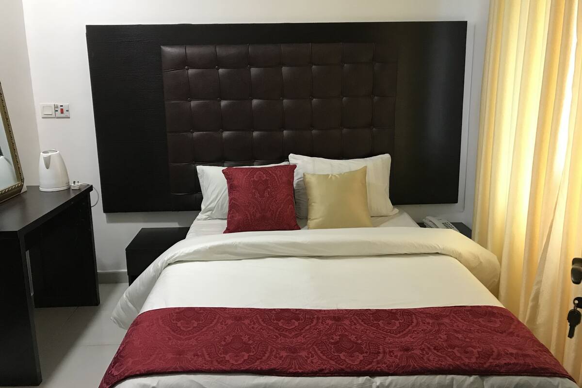A hotel room with a large dark headboard, a bed with white and red bedding, a bedside table with a lamp, and a curtain on the window.