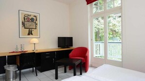A room with a bed, a desk with a chair, a television, and a window with a view of trees. - Art Hotel Weimar (Weimar)