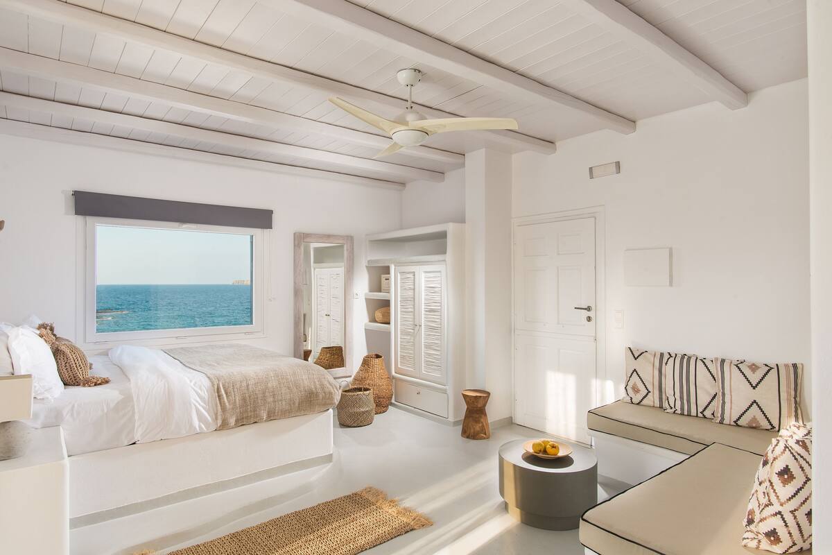 A bedroom with a bed, a window with a view of the sea, a ceiling fan, and a small round table with a bowl of fruit.