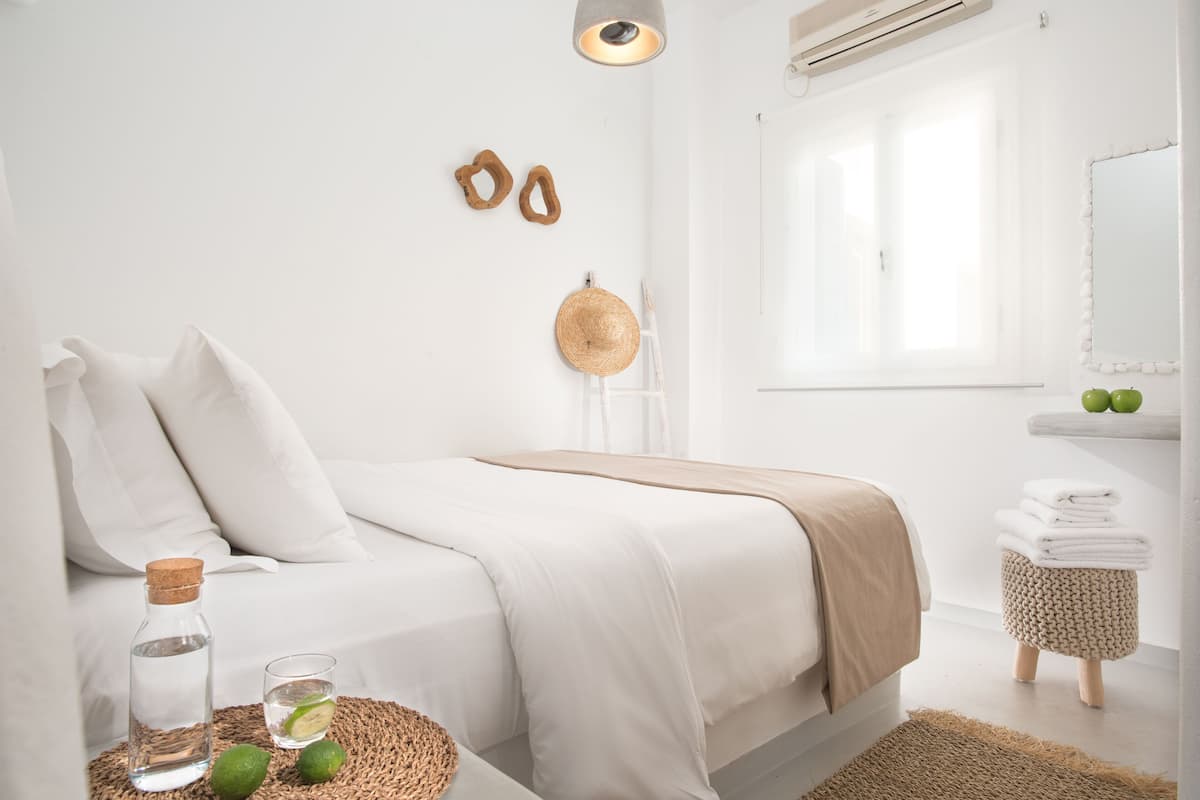 A minimalist bedroom with a bed, a bedside table with a bottle and lime, a woven mat, a small stool, and a window with a view.