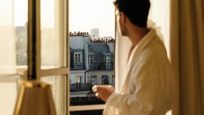 A person in a robe standing by an open window with a view of buildings. - 9Confidentiel (Paris)
