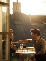 A couple dining at an outdoor table with a view of a building's rooftop and a clock tower in the background.