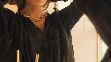 A person in a black dress with a gold chain necklace standing in front of a bathroom mirror.