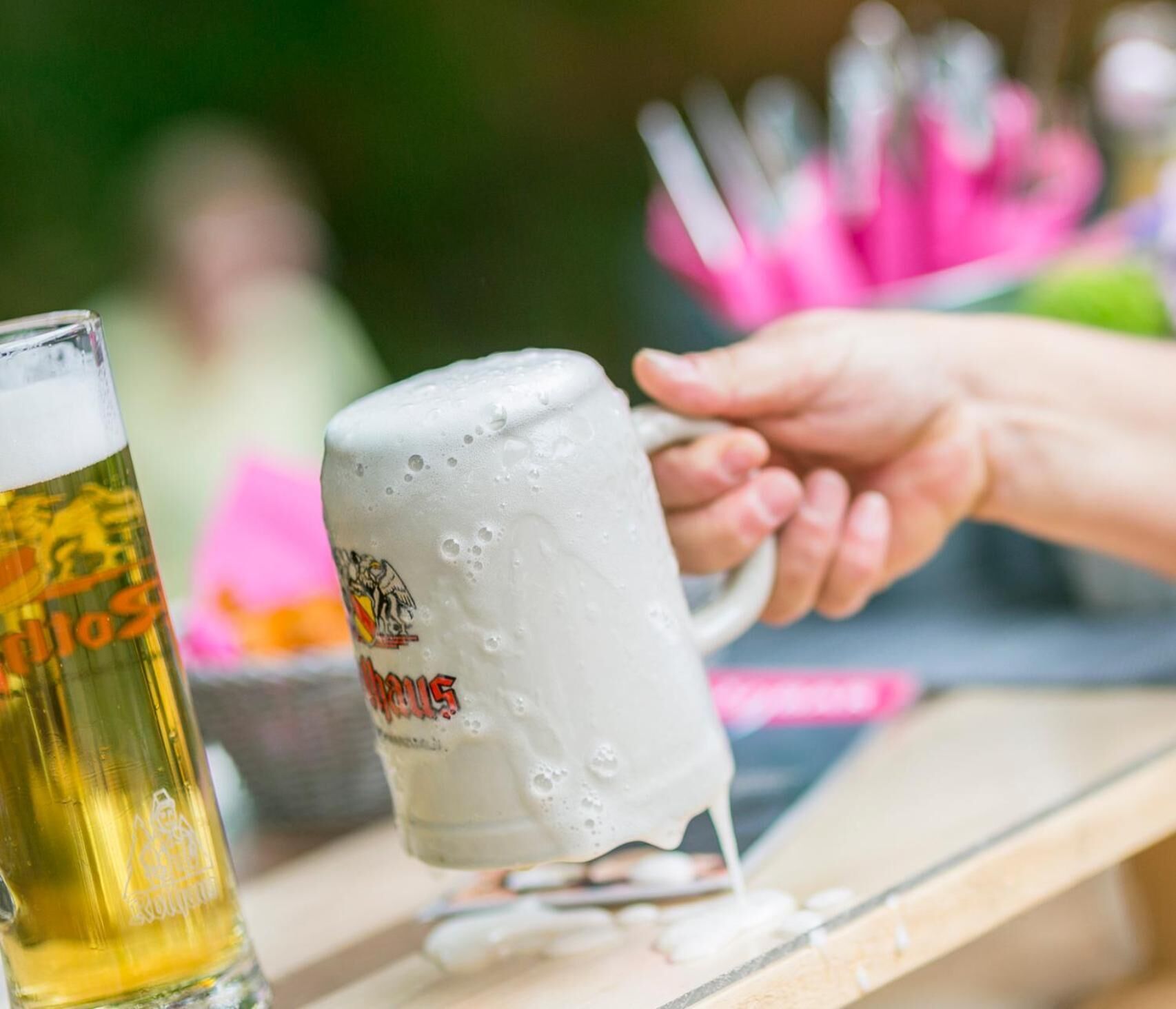 Une main tenant une chope de bière mousseuse ornée d’un logo, à côté d’un verre de bière.