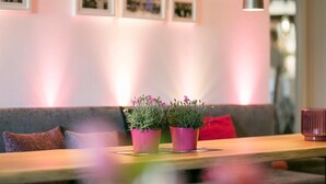 A seating area with a wooden table, two potted plants, a red cushion, and wall-mounted pictures. - Hotel Imbery (Hinterzarten)