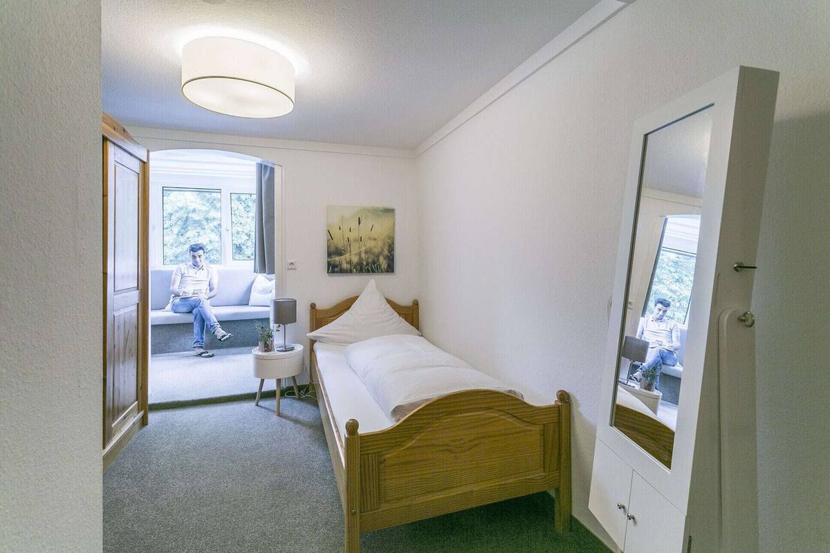 Un dormitorio con una cama, una lámpara de noche, un cuadro, un espejo y vistas a un balcón donde se sienta una persona.