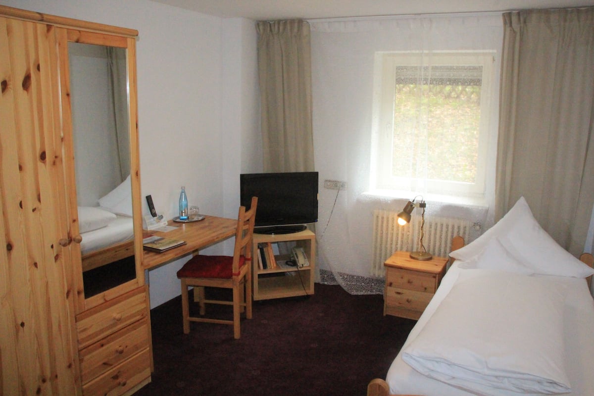 Una habitación con una cama, un escritorio con televisor, una silla, una mesita de noche con lámpara y una ventana con cortinas.