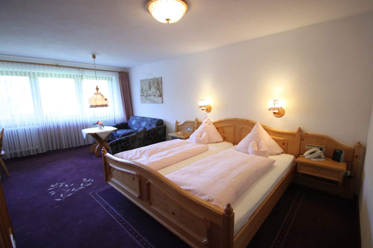 Una habitación de hotel con una cama de madera, una mesita pequeña, una silla, una mesita de noche y una lámpara.