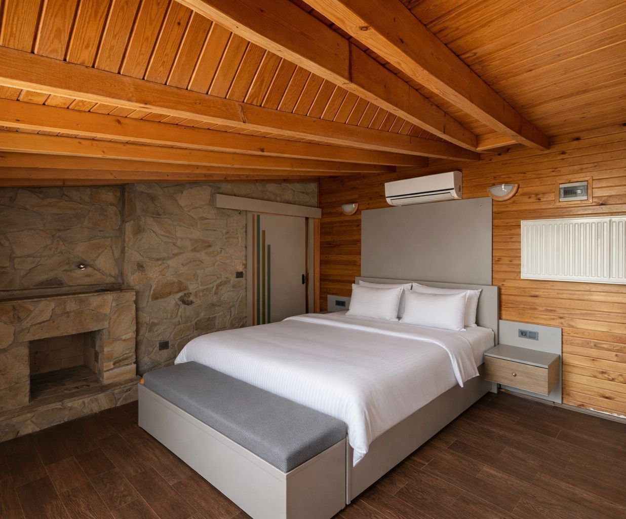 A bedroom with a stone fireplace, a bed with a grey bench, wooden ceiling beams, and a white radiator.