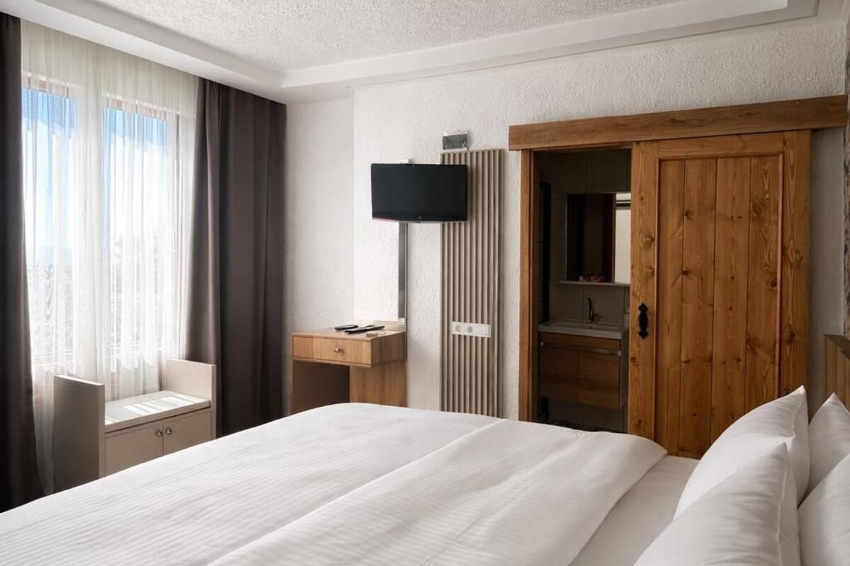 Une chambre d’hôtel avec un grand lit, une armoire en bois, un bureau avec une chaise, une télévision et une fenêtre avec des rideaux.