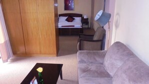 A living room with a sofa, a coffee table with a vase, a bed with a headboard, a chair, and a lamp. - Pacific Hotel (Addis Ababa)