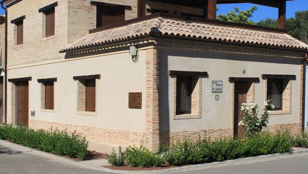 A two-story building with a tiled roof and brick walls, featuring a covered patio area and a small garden with shrubs.