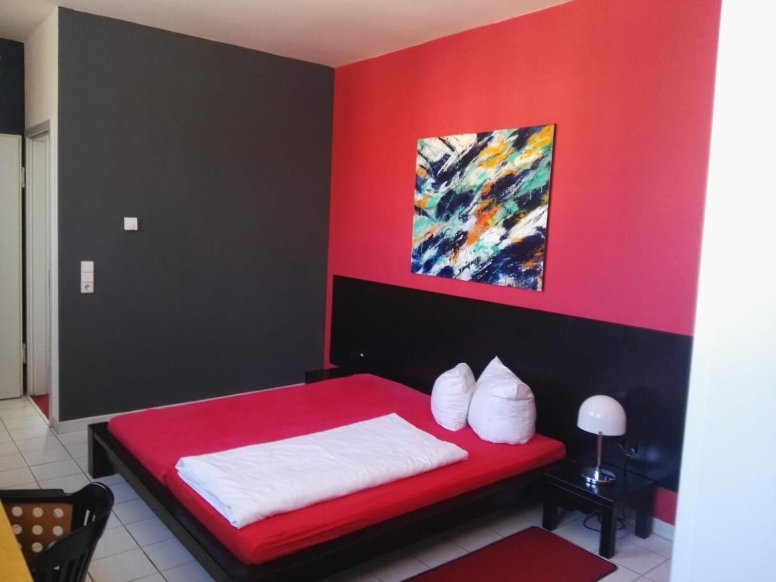 Un dormitorio con una colcha roja, un almohadón blanco y un cuadro abstracto de colores en la pared.