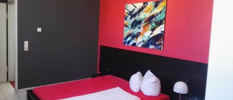 Un dormitorio con una colcha roja, un almohadón blanco y un cuadro abstracto de colores en la pared.