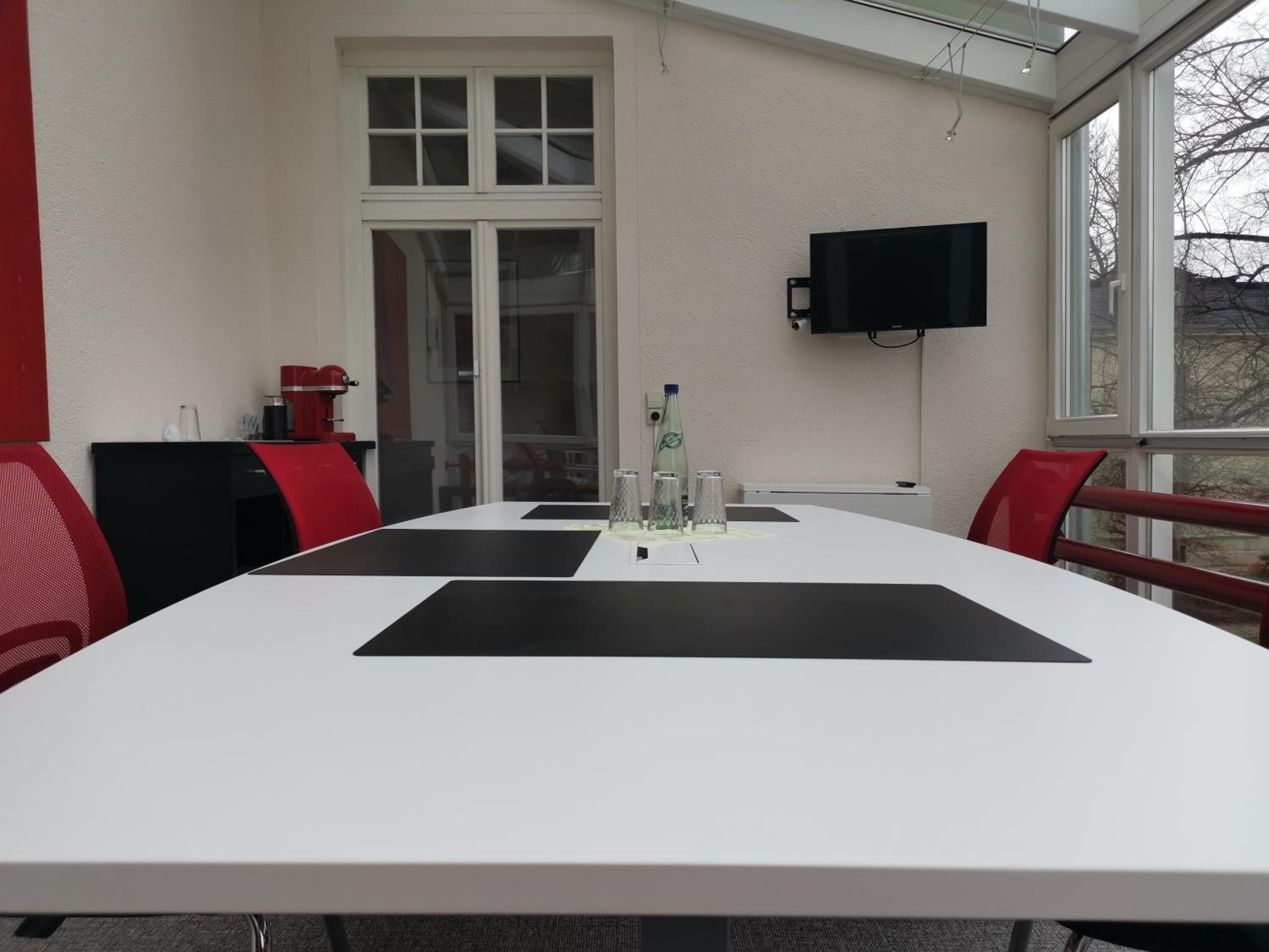 A conference room with a white table, red chairs, a TV on the wall, and a door with glass panels.