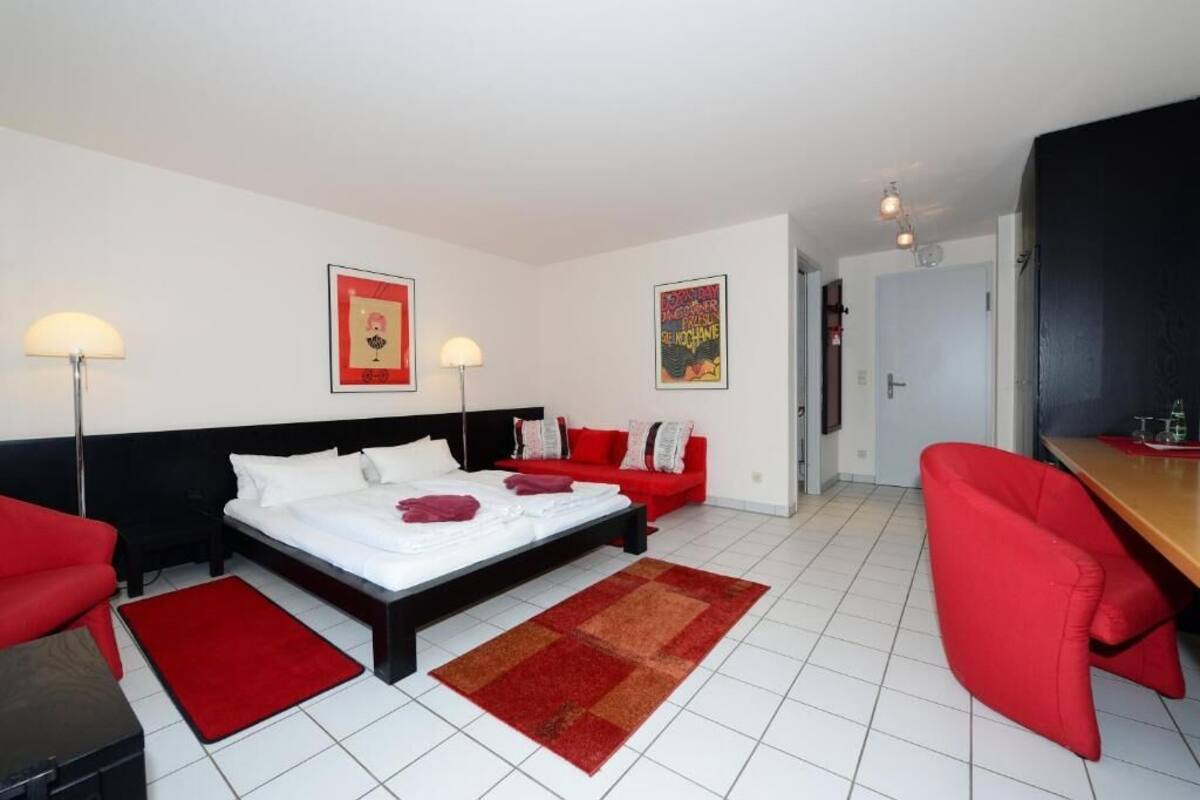 A hotel room with a bed, a red sofa, a red armchair, a desk, and two wall-mounted lamps.