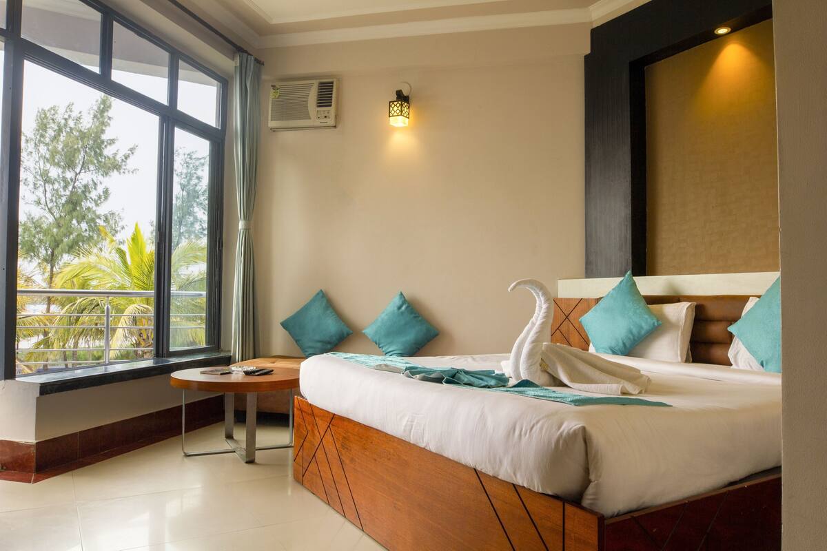 Een hotelkamer met een groot bed, een klein tafeltje, een raam met uitzicht op palmbomen en een wandairconditioner.
