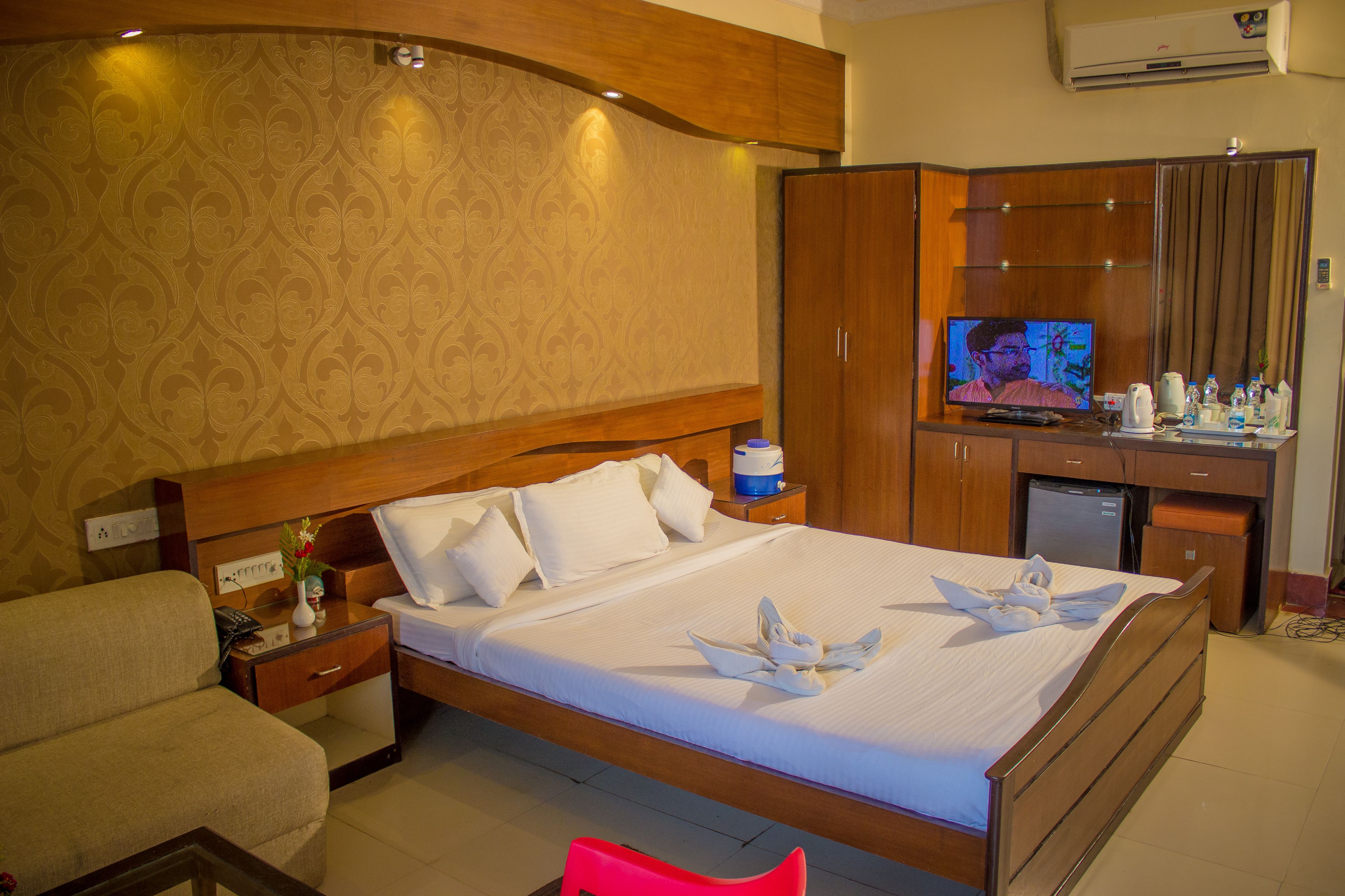 A hotel room with a large bed, a wooden wardrobe, a television, and a small desk with a kettle and bottles.