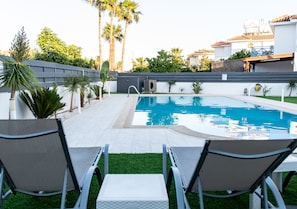 A swimming pool with two lounge chairs in the foreground, surrounded by palm trees and a white wall. - Long Beach Gardens 1 (Oroklini)