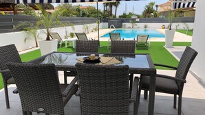 Une terrasse dotée d’une table en verre et de chaises en osier, offrant une vue sur une piscine et une pelouse verdoyante.