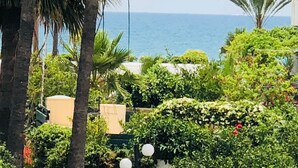 Un jardin luxuriant, avec des palmiers et un feuillage dense, une vue dégagée sur la mer et un bâtiment à la façade beige.