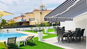 Une piscine avec des chaises longues, un coin repas avec des chaises et une terrasse couverte agrémentée d’auvents à rayures.