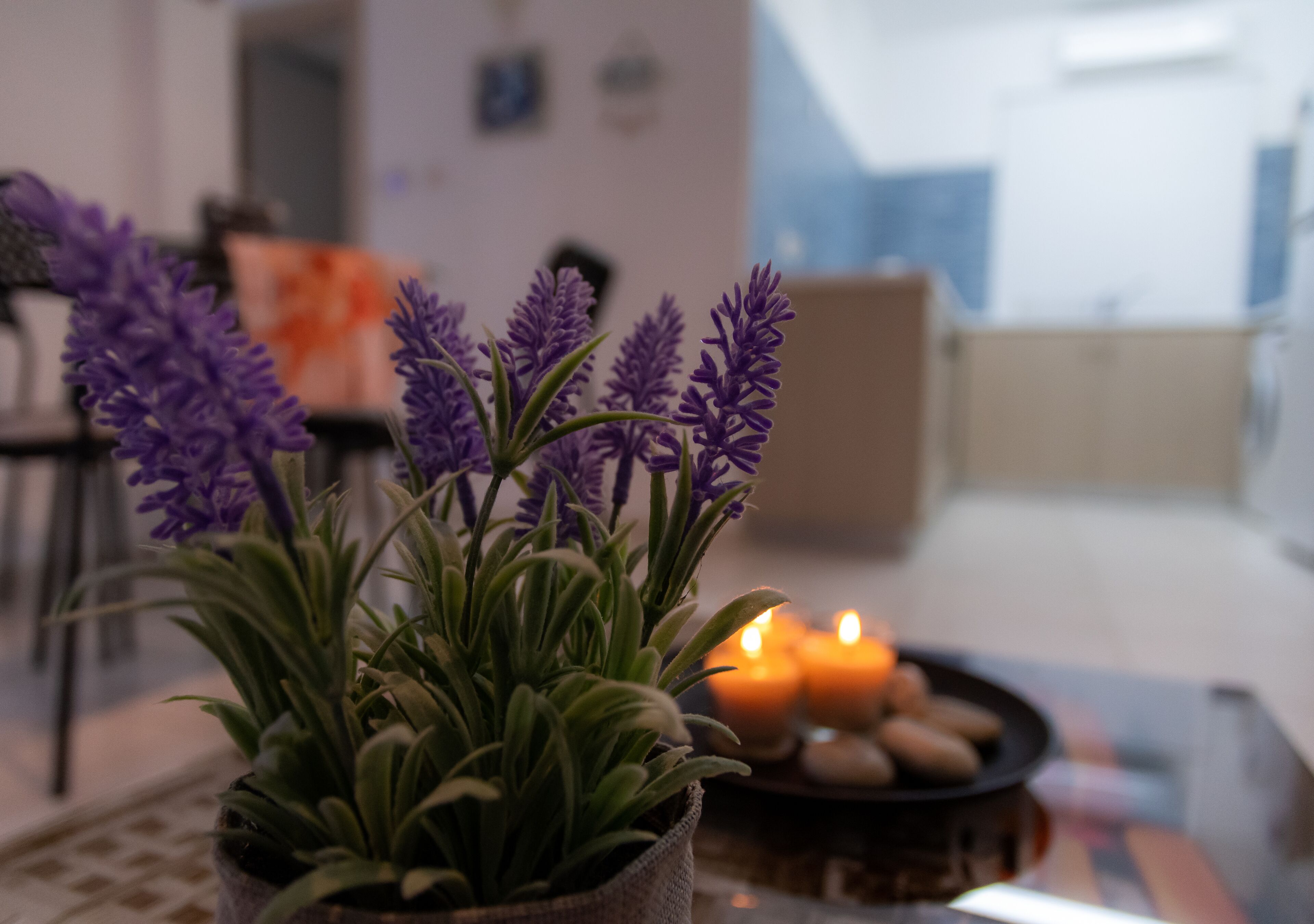 Una planta en maceta con flores moradas sobre una mesa, velas y un cuenco de piedras en un salón.