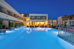 A modern hotel with a large indoor swimming pool, multiple balconies, and lounge areas with umbrellas. - White Olive Premium Laganas - All Inclusive (Zakynthos)