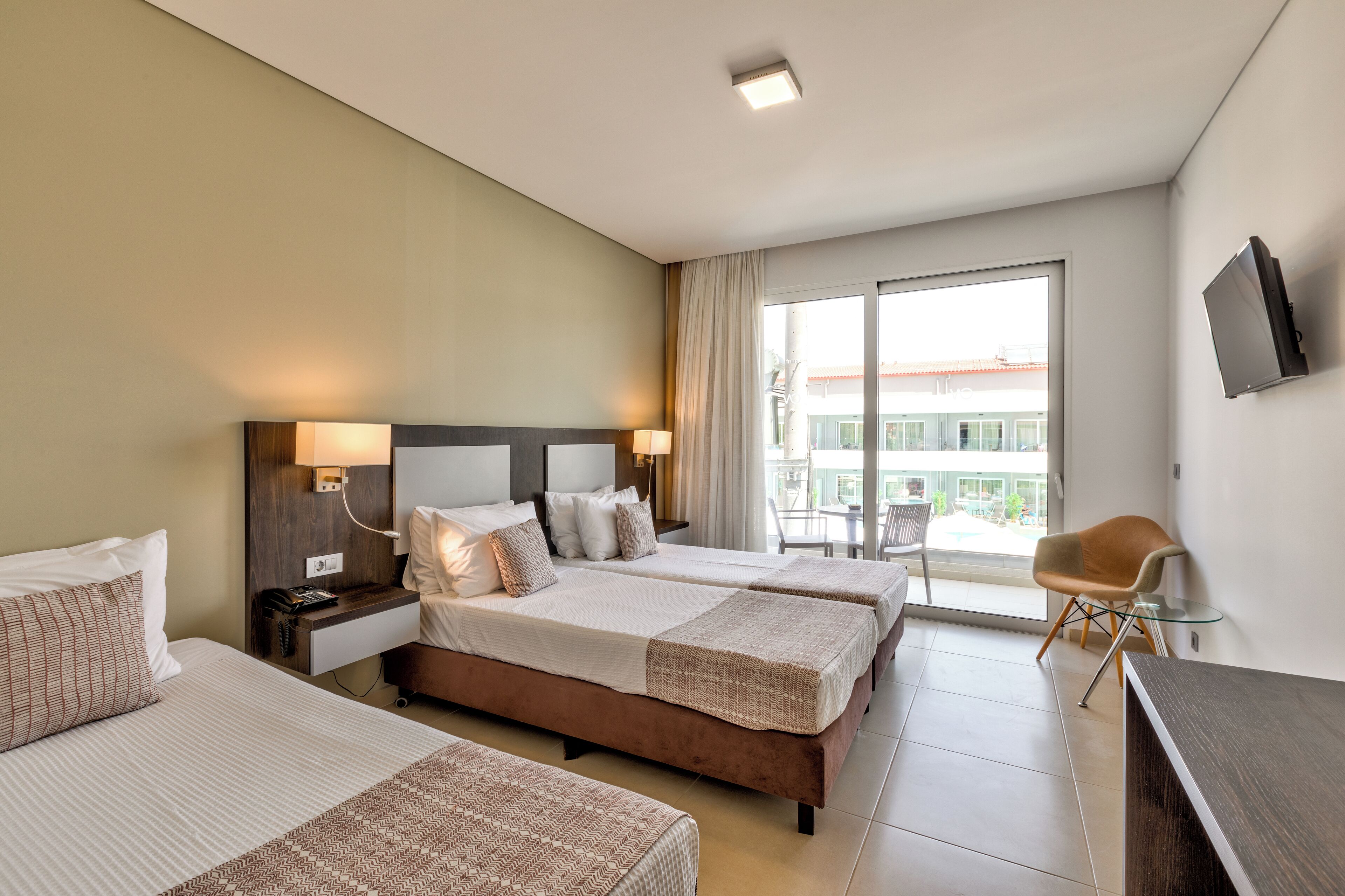 A hotel room with two beds, a desk, a chair, and a large window with a view of buildings.
