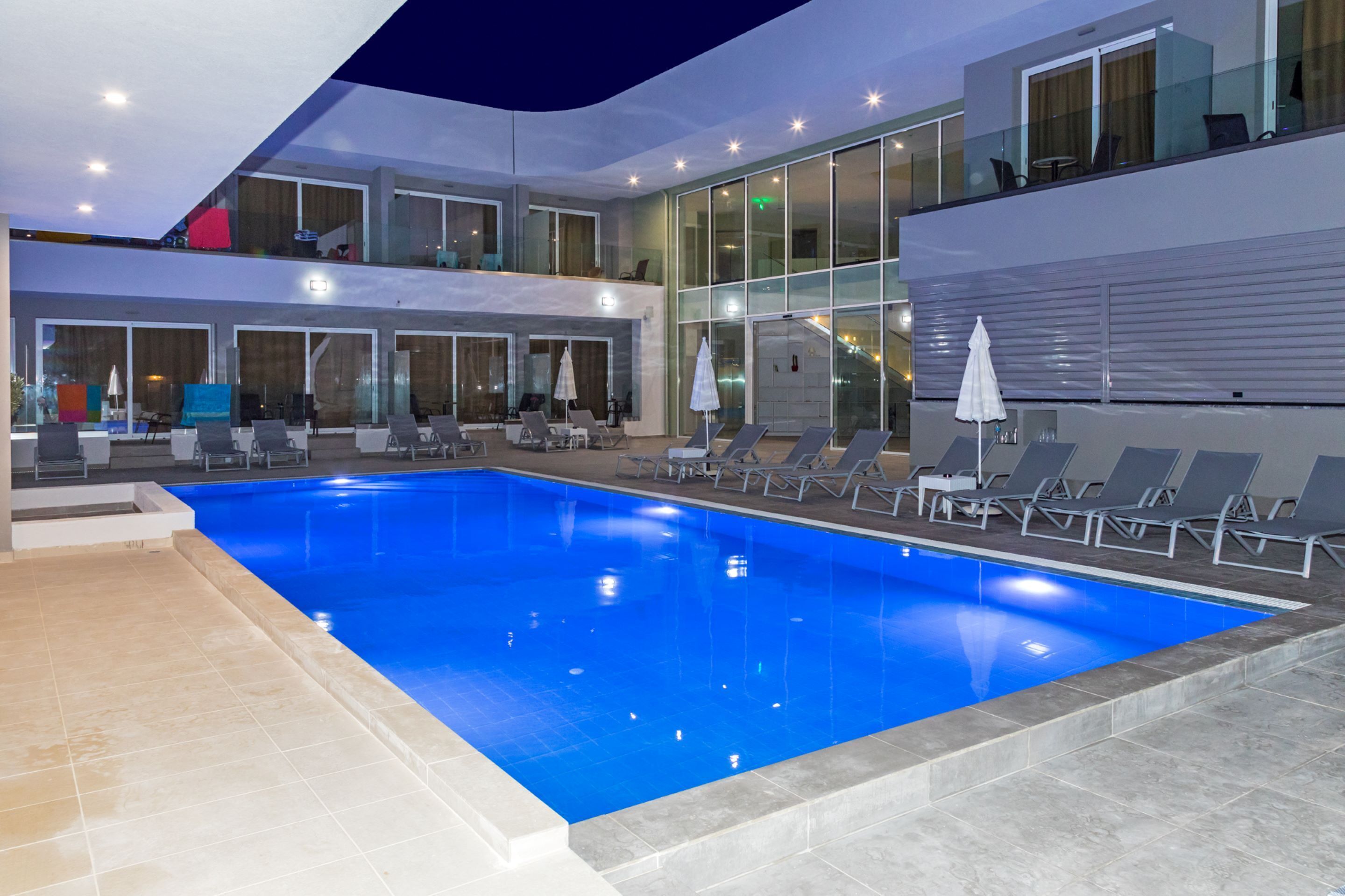 Una piscina interior moderna con sillones de descanso y sombrillas, ventanas amplias y un balcón de varios niveles.