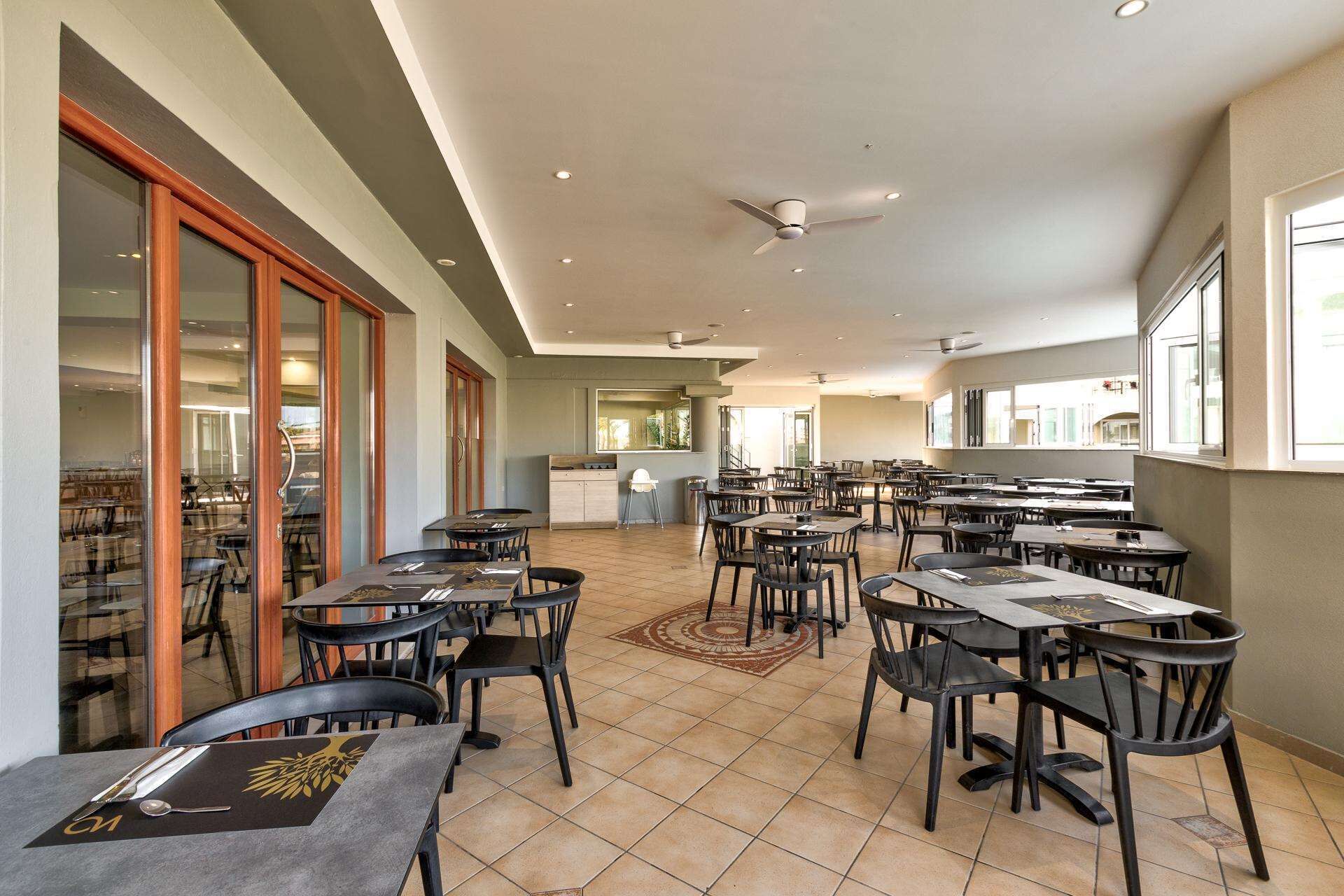A spacious indoor dining area with multiple tables and chairs, a kitchen in the background, and large windows allowing natural light.