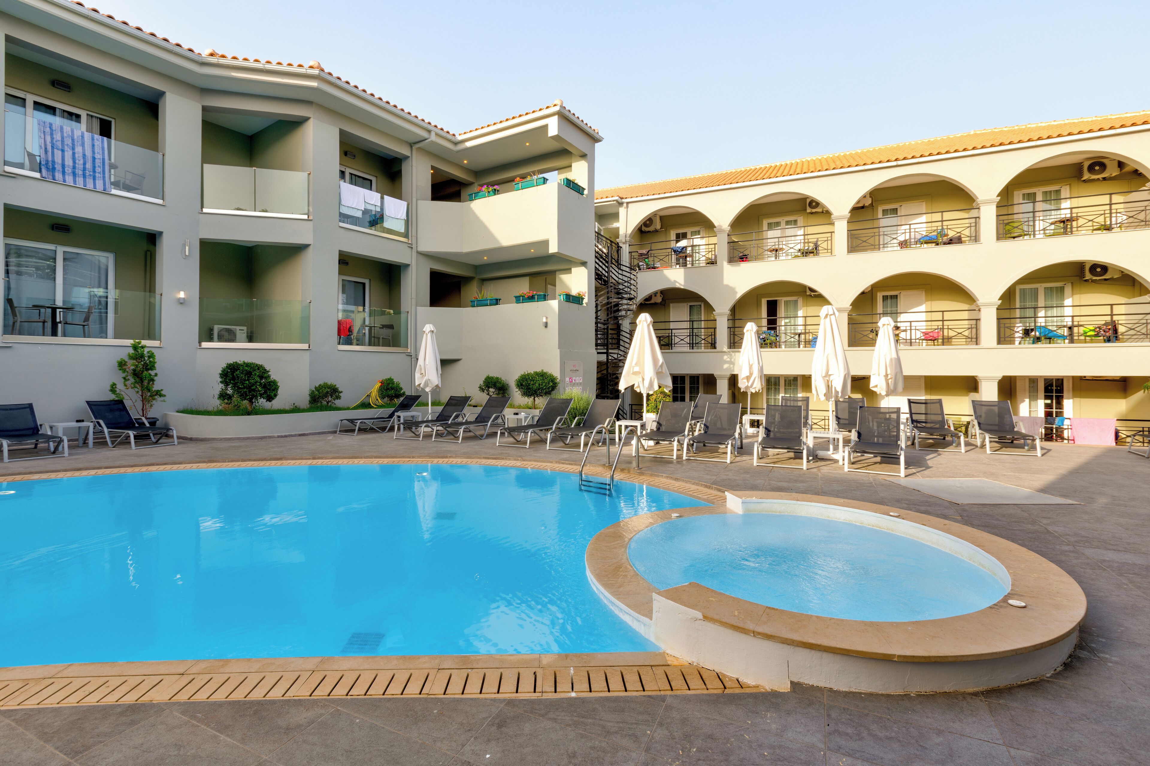 Una piscina con lettini e ombrelloni, circondata da un edificio a più piani con balconi e zone salotto all'aperto.