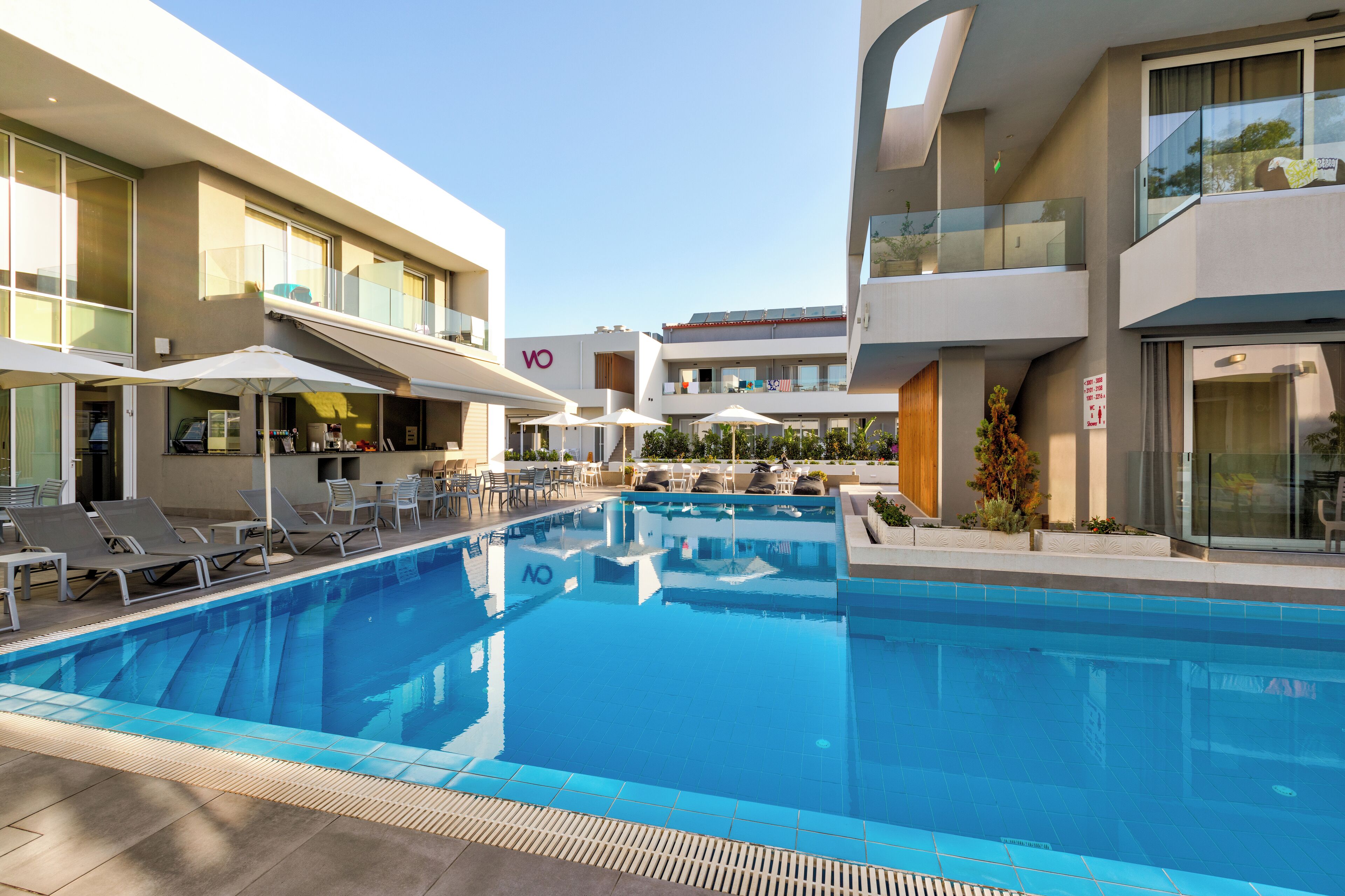 A modern hotel with a swimming pool, outdoor seating, and a clear sky.
