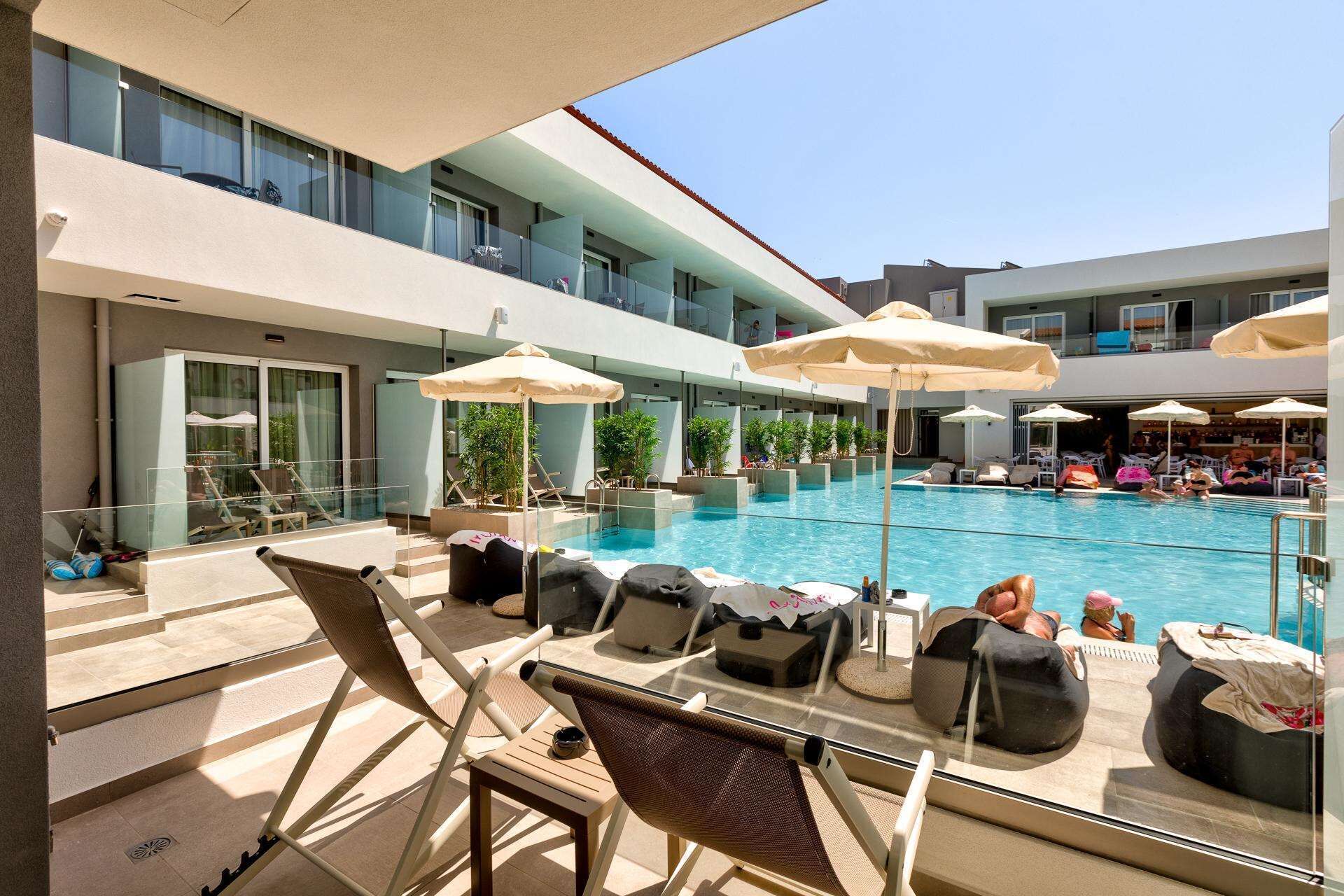 A poolside area with lounge chairs and umbrellas, a clear view of the pool, and a building with multiple balconies.
