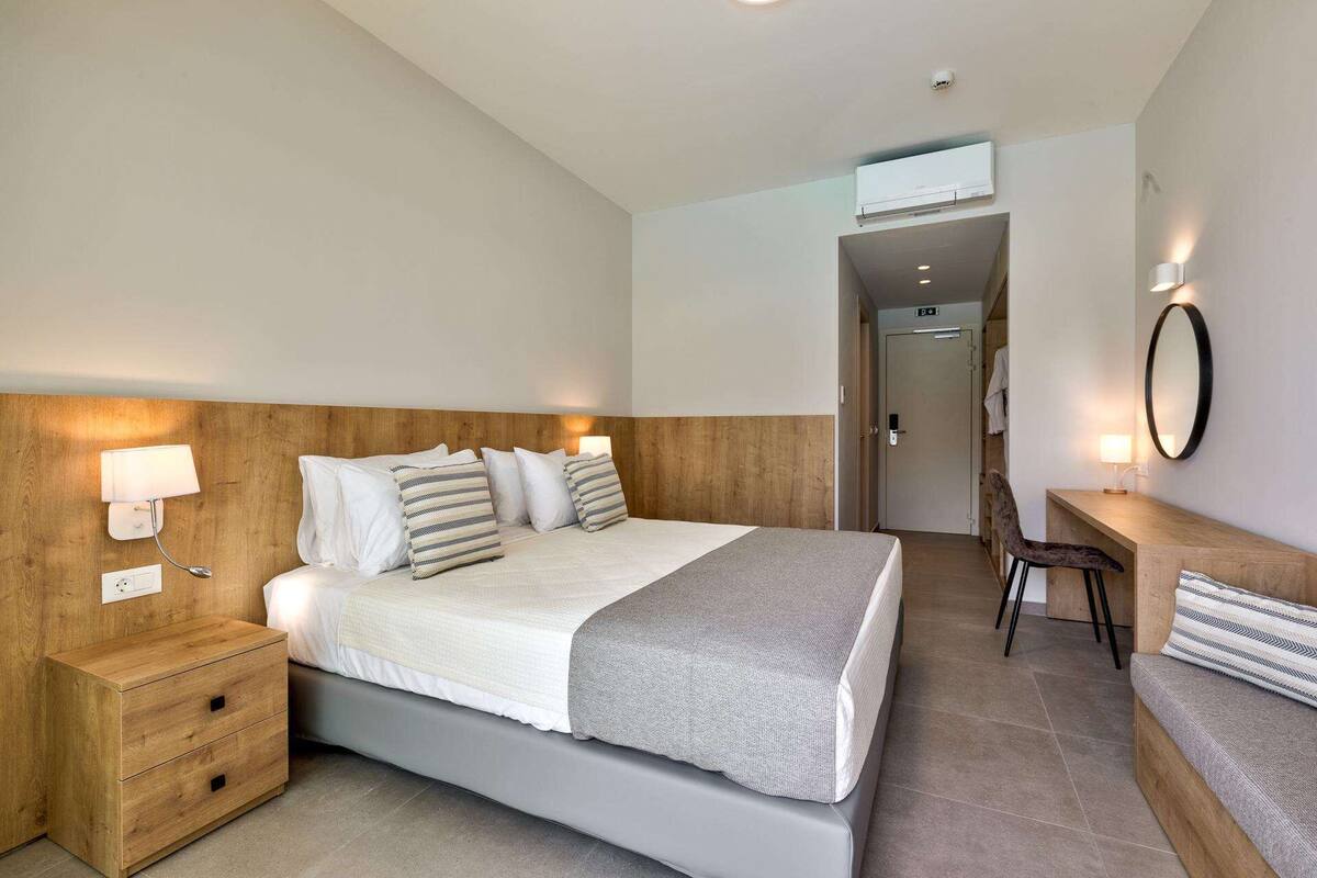 A modern hotel room with a wooden headboard, a large bed with multiple pillows, a bedside table with a lamp, a desk with a chair, and a mirror on the wall.