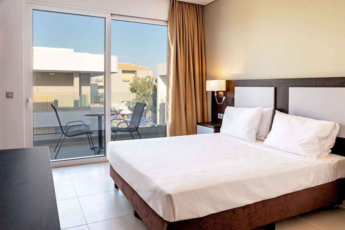 A hotel room with a bed, a nightstand, a lamp, a view of a pool and lounge chairs through a sliding door.