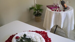 A neatly made bed with a white towel and rose petals, a small potted plant, and a table with a bottle of champagne and glasses. - Tropical Stay Accommodations (Tagbilaran)