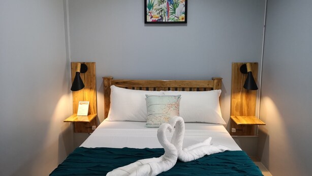 A neatly made bed with a teal blanket and a white towel arranged in a swan shape, flanked by two bedside lamps and a framed tropical artwork above. - Tropical Stay Accommodations (Tagbilaran)