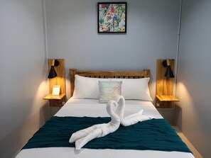 Een net opgemaakt bed met een blauwgroene deken en een handdoek gevouwen in de vorm van een zwaan, met aan weerszijden nachtlampjes en erboven een kunstwerk met een tropisch thema.