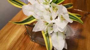 A clear glass vase with white flowers and green-striped leaves on a wooden surface. - Tropical Stay Accommodations (Tagbilaran)