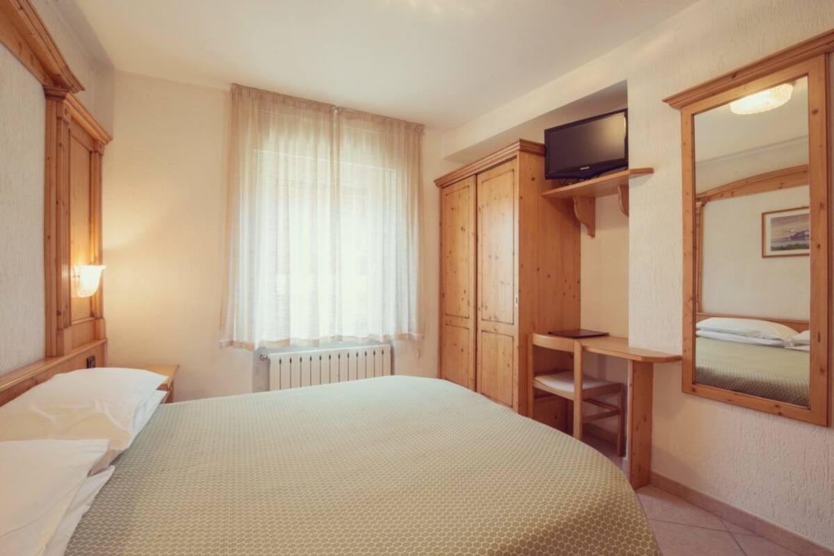 Un dormitorio con cama, un armario de madera, un escritorio con silla, televisión en un estante y ventana con cortinas.