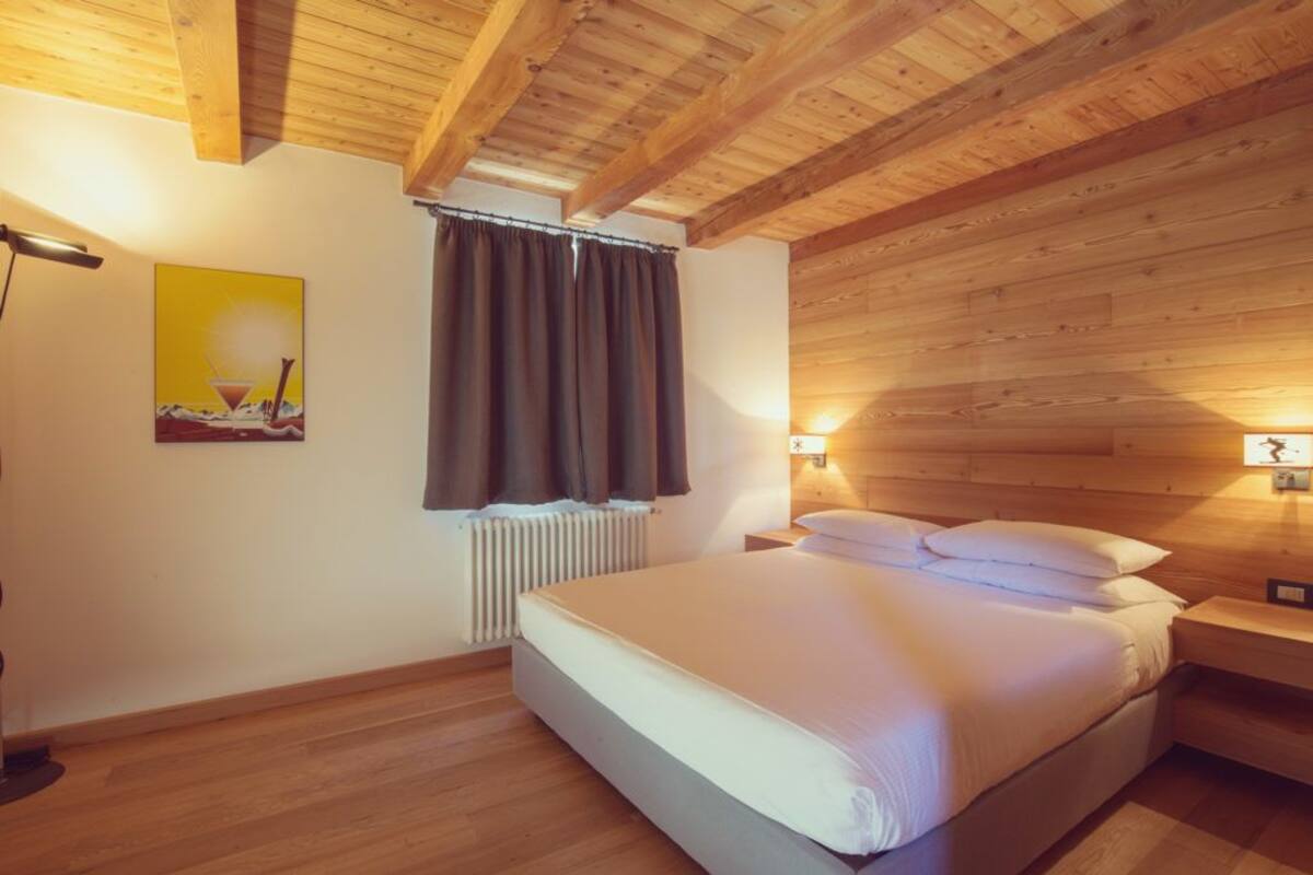 Un dormitorio con techo de madera, una cama con ropa blanca, una mesita de noche, una lámpara, una ventana con cortinas y un radiador.