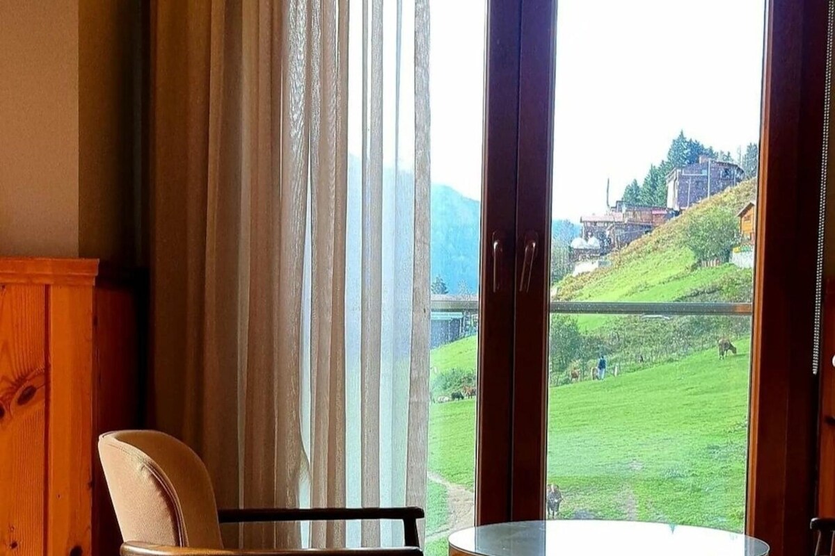 A room with a bed, a chair, a table, and a large window with a view of a green landscape and buildings.