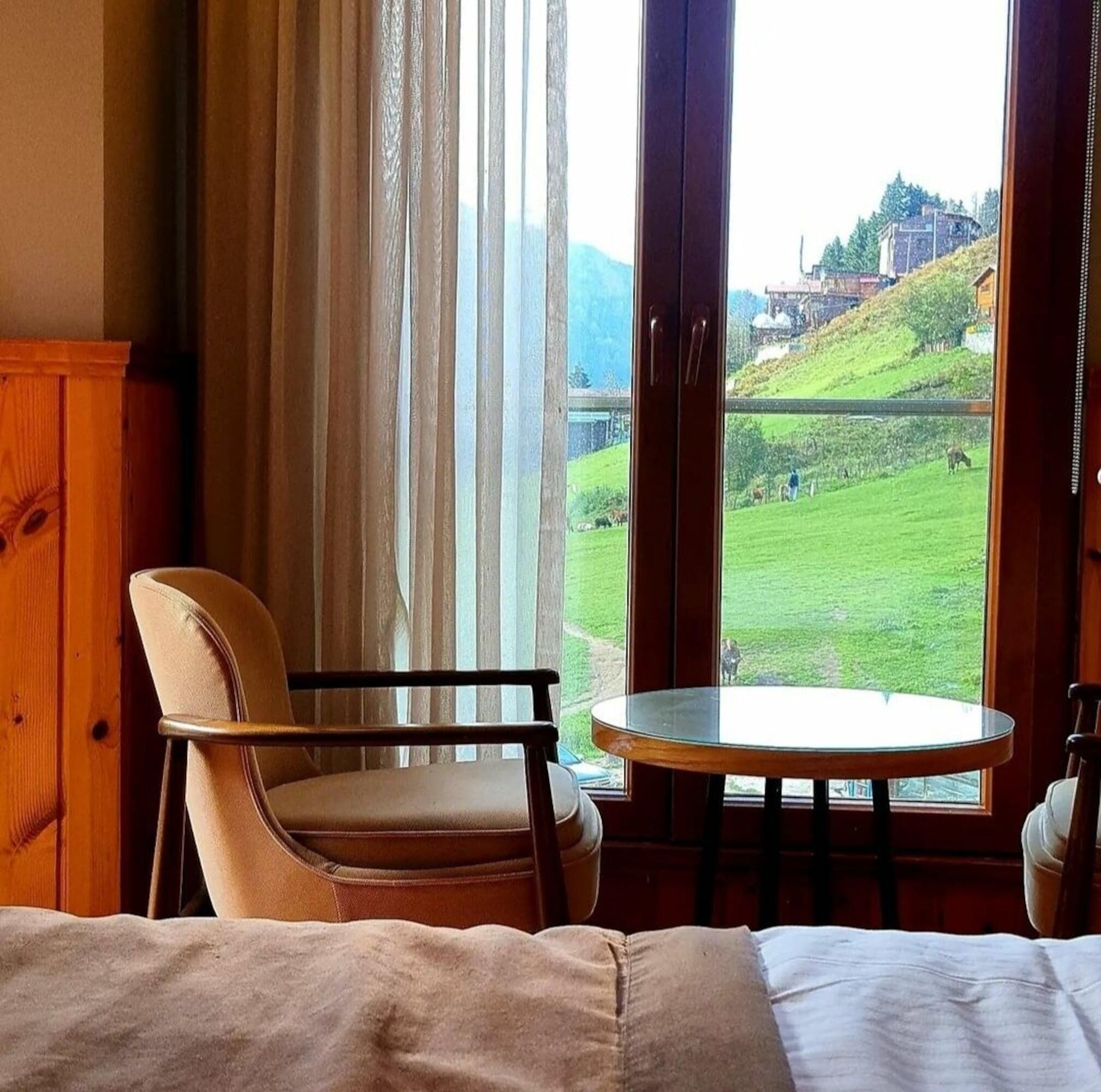 Una habitación con cama, una silla, una mesa y un ventanal con vistas a un paisaje verde y edificios.