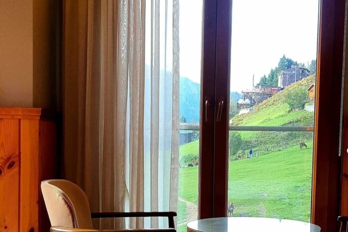 A room with a bed, a chair, a table, and a large window with a view of a green landscape and buildings.